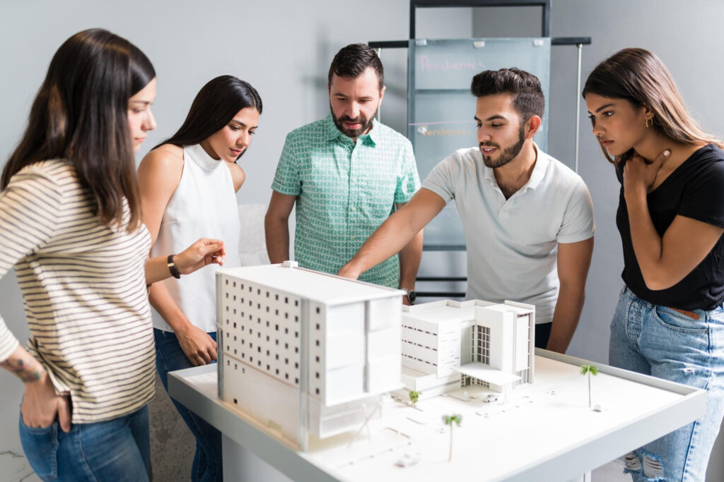 Five people standing around a model building, all looking closely as one of the five, points at a fine detail within the model.